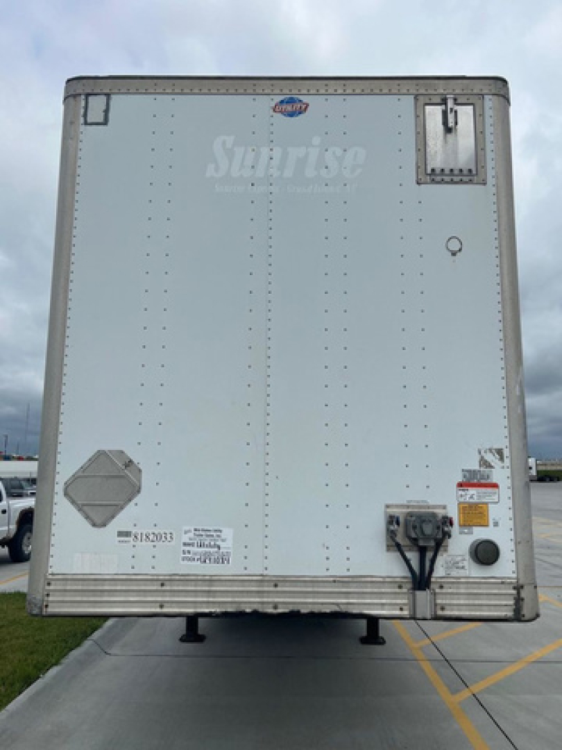 Back of a silver utility trailer with a small vent door, bumper hitch, and branding stamp. Sun logo near top, label stickers bottom left, rear wiring and safety decals visible.
