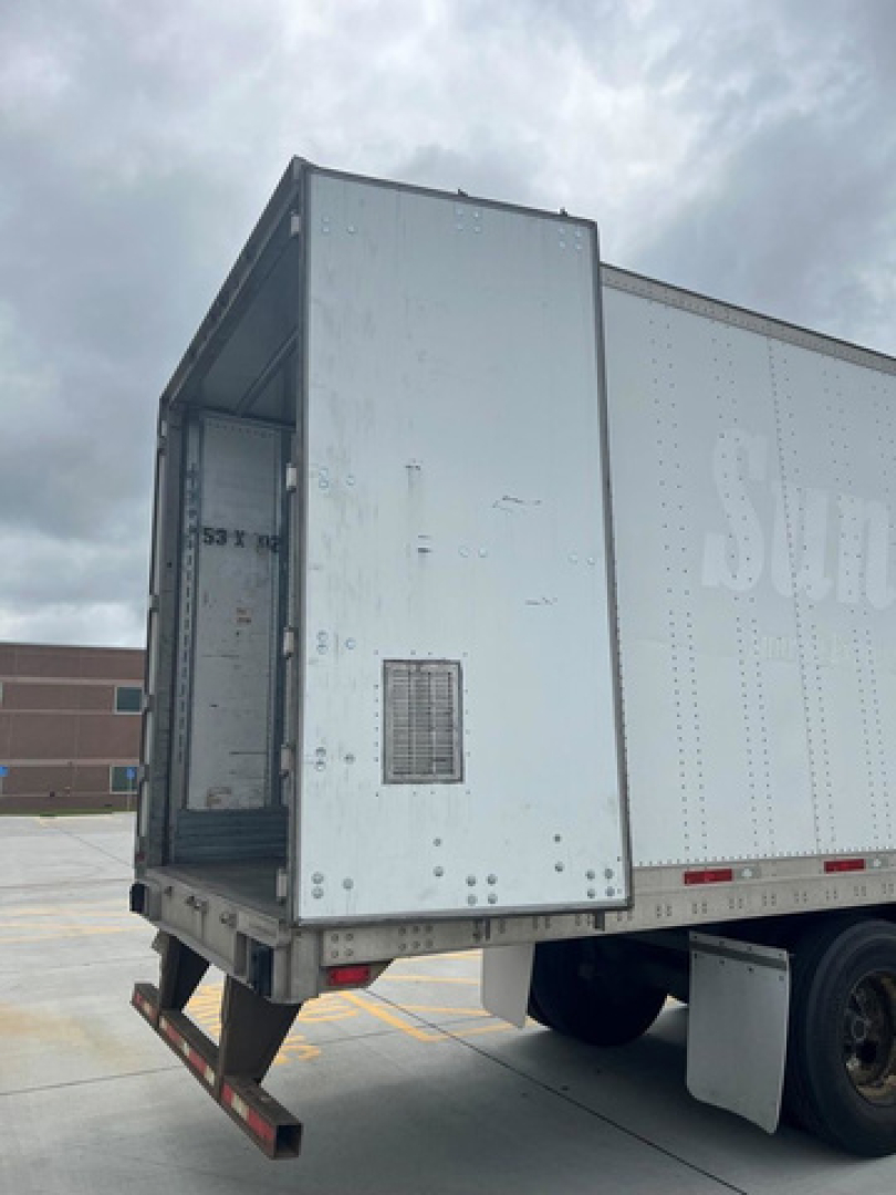 Trailer rear showing open roll-up door and loading ramp, silver exterior with riveted panels.