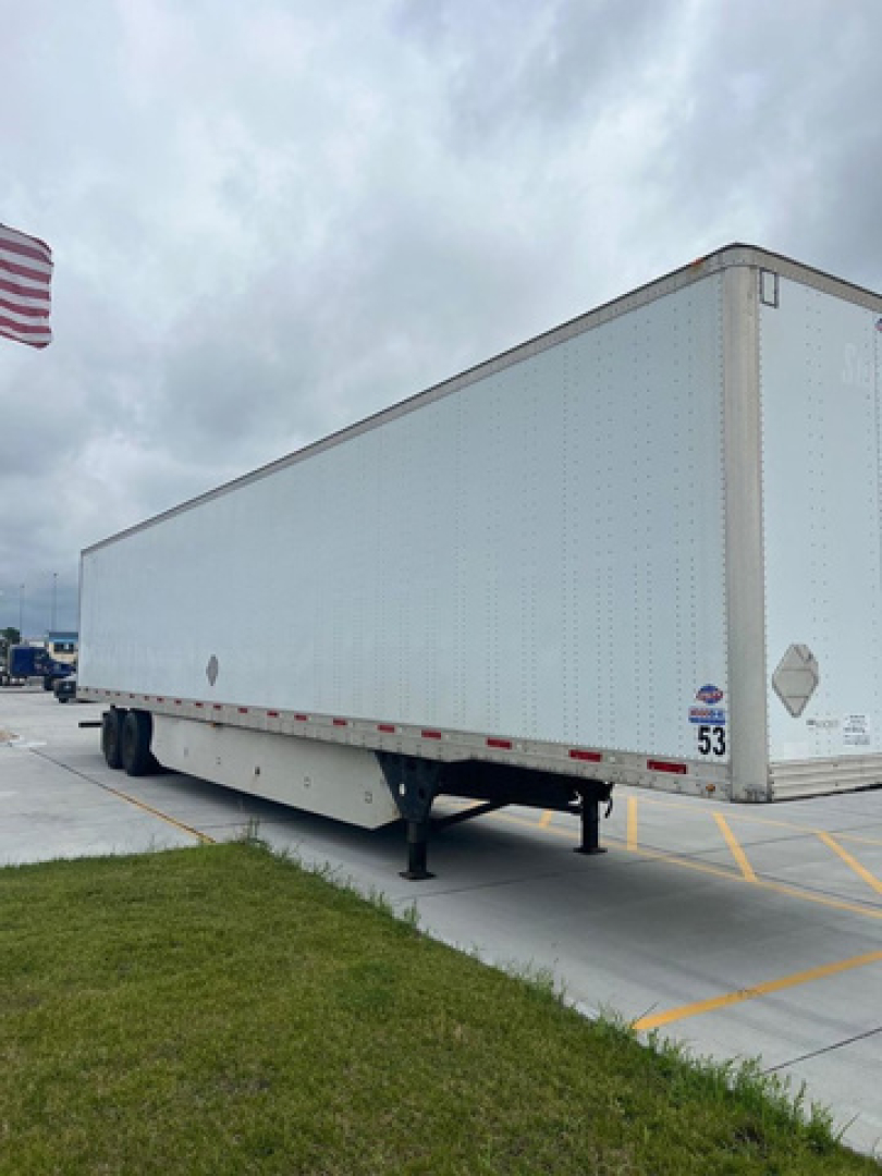 Utility van trailer with white exterior, parked on pavement; visible front landing gear and side stickers; US flag in background.