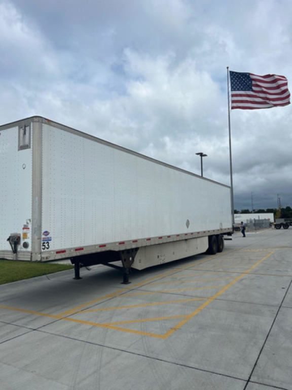 Utility van trailer parked at a lot with an American flag, viewed from the rear left side, showing a long white box trailer on level ground.