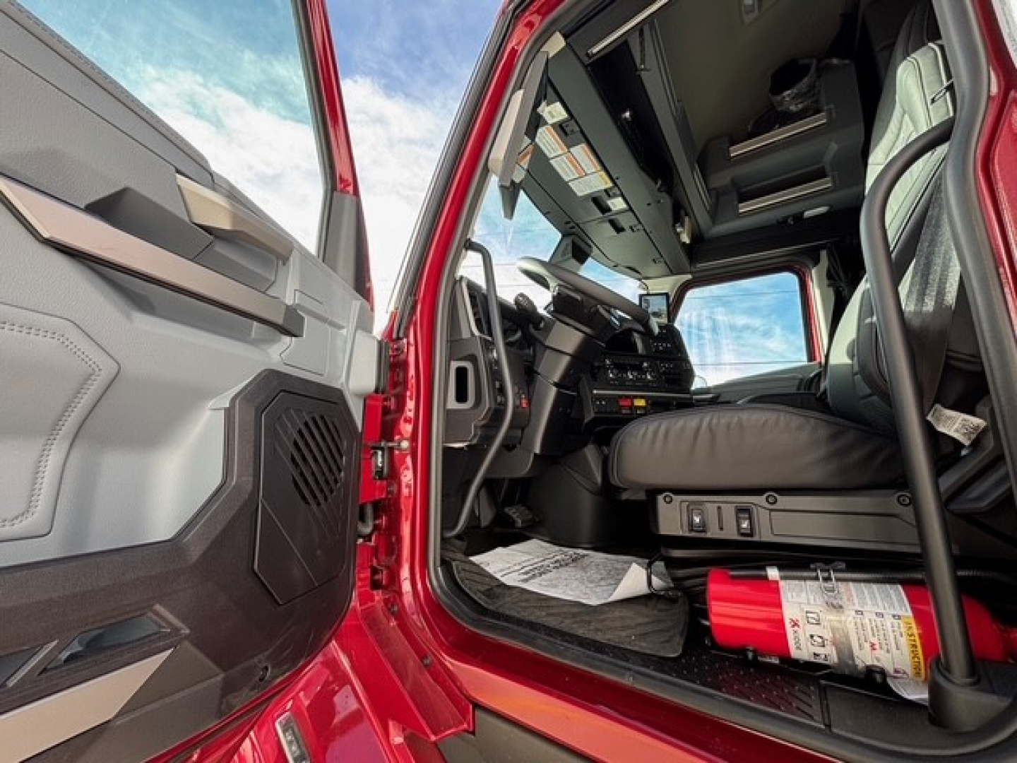 Cab interior of a 2026 Mack PR64T76H truck showing driver seat and dashboard with open door; red exterior visible near door sill.
