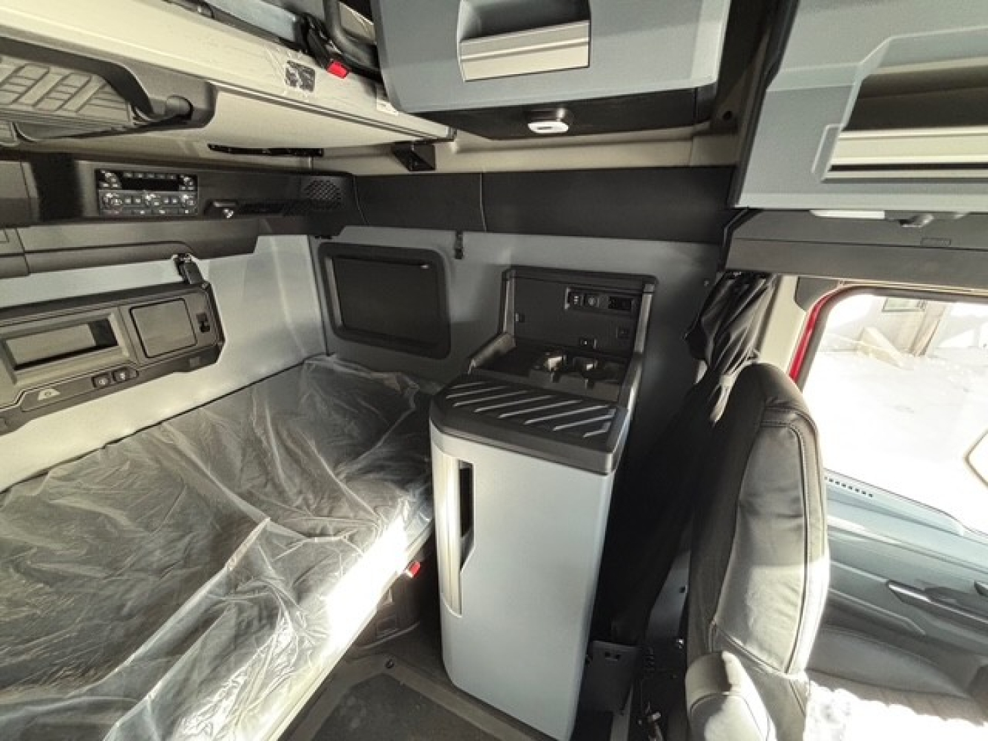 Interior of a new 2026 Mack PR64T76H truck cab with sleeper area, showing bunk bed area covered in plastic, overhead storage, and a center console with controls.