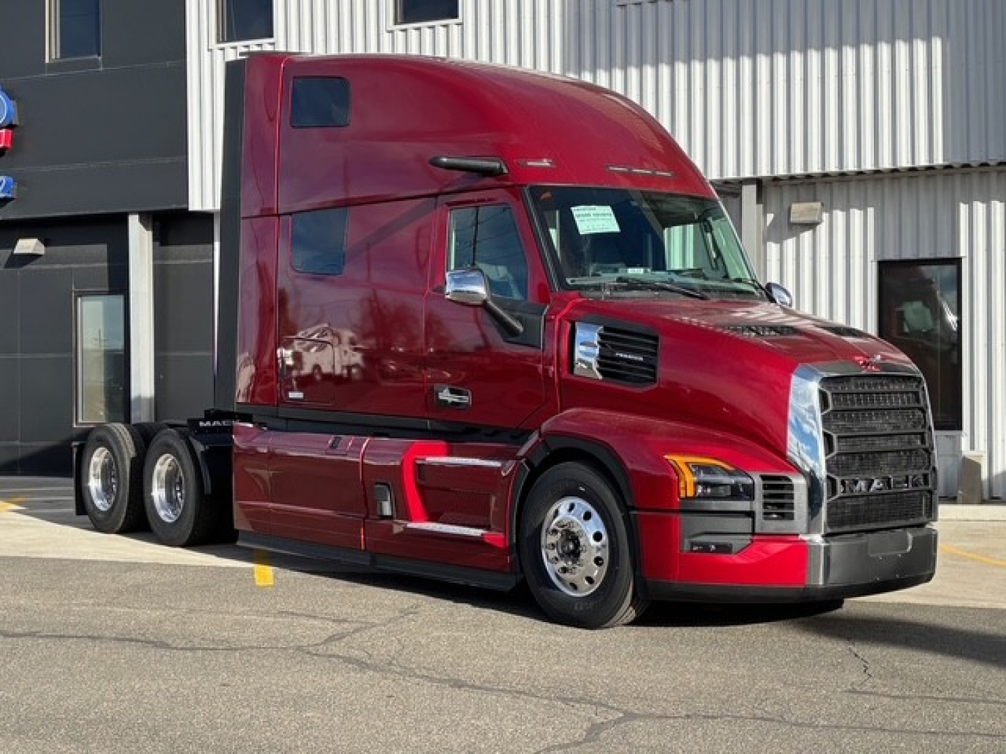 Truck cab with red Mack PR64T76H sleeper tractor, side view showing front grille and fuel tanks, no text visible.