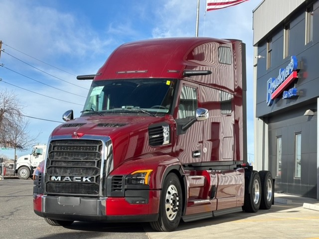 Truck bay showing a red 2026 Mack PR64T76H tractor with triple rear axles parked outside a dealership.