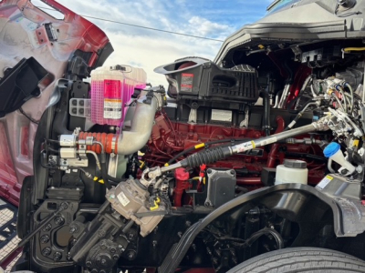 Mack PR64T76H engine bay showing red diesel engine with various hoses, pink coolant reservoir, and wiring harnesses in a conventional sleeper truck.