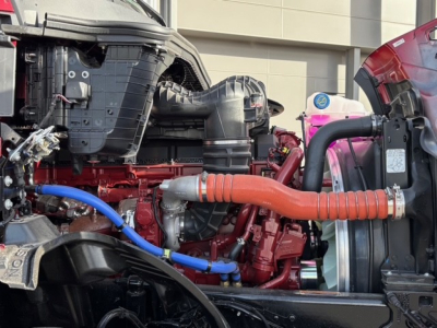 Engine bay of 2026 Mack PR64T76H showing red engine block with orange coolant hose and blue hose, radiator and intake components visible.