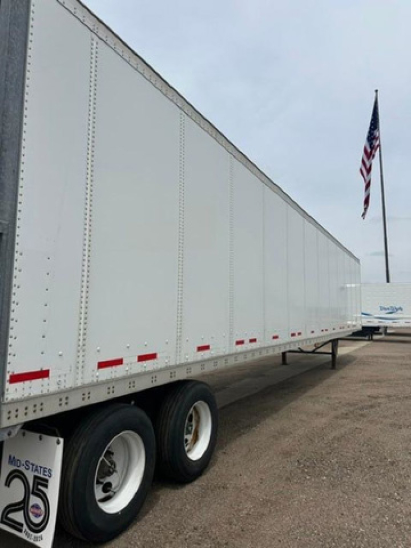 Trailer side view showing a long white 2016 Wabash van trailer with dual rear wheels and reflective red markers along the lower edge; US flag in background.