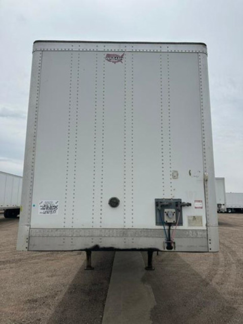 Rear view of a white Wabash van trailer with stabilizer legs visible, rear doors closed, with a hitch and utility box on the lower right; small stickers near left door edge.