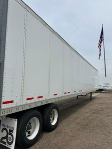 Trailer side view showing a long white 2016 Wabash van trailer with dual rear wheels and reflective red markers along the lower edge; US flag in background.