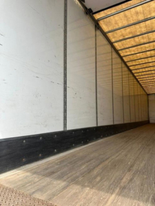 Interior side of a trailer with wooden floor and white walls, showing a long empty cargo area. There are no legible labels or text visible in the image.