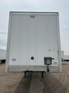 Rear view of a white Wabash van trailer with stabilizer legs visible, rear doors closed, with a hitch and utility box on the lower right; small stickers near left door edge.