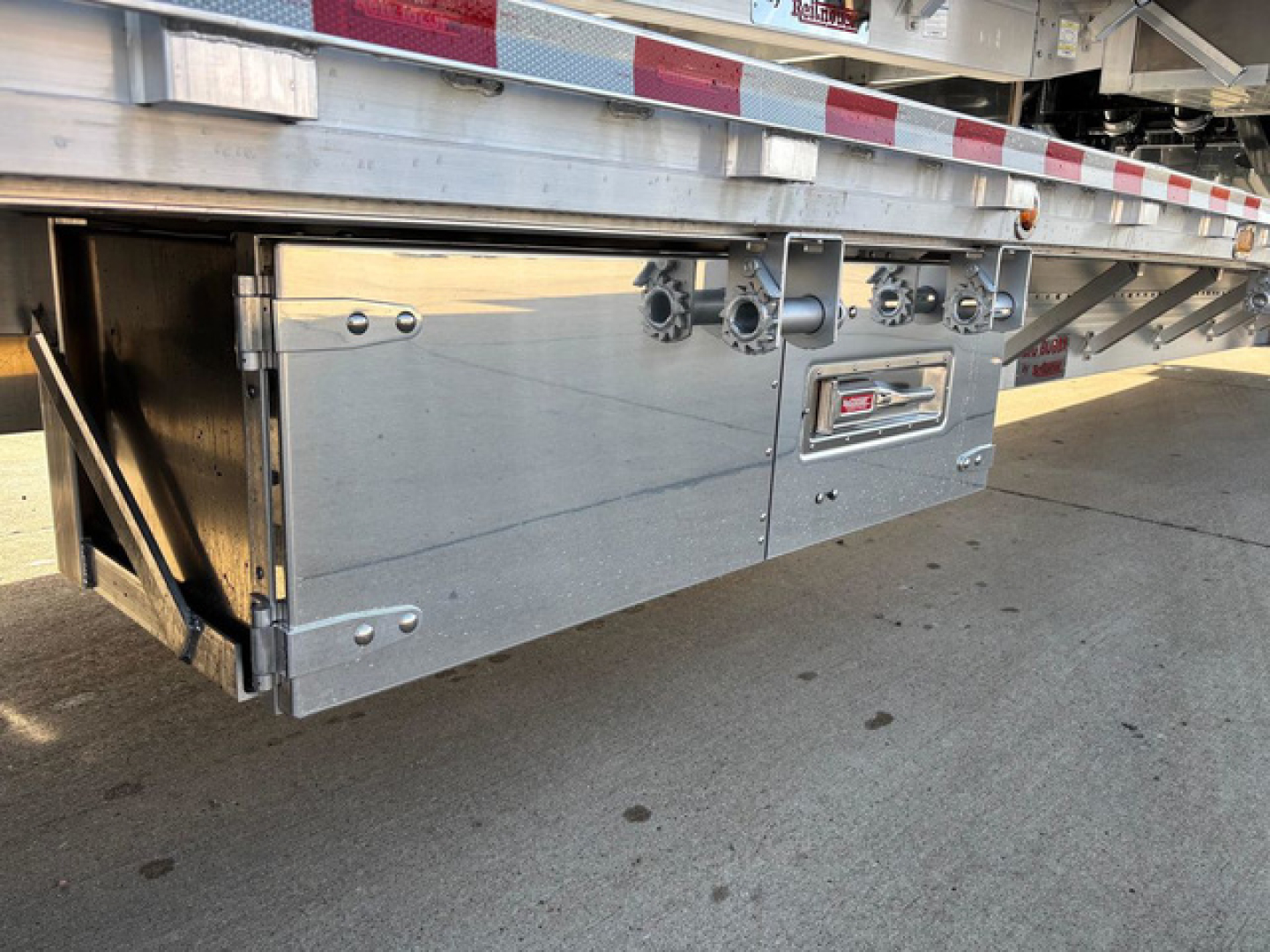 Dropdeck trailer side with metal access panels and hinge details; visible are bolted box compartments and a small label area.