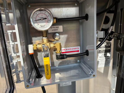 Trailer toolbox interior showing pressure gauge, brass valve assembly, yellow tag, and label 'AIR BAGS EMPTY' on the side; gear gauge reading and plumbing visible.