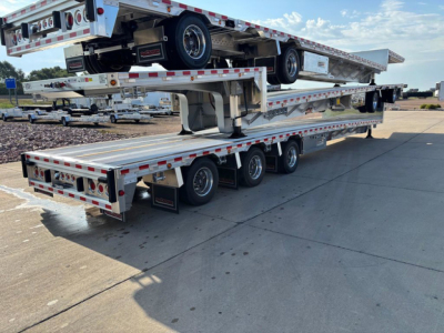Drop deck trailer stacked with extendable flatbeds and rear ramps, showing multiple axles and reflective tape along the sides.