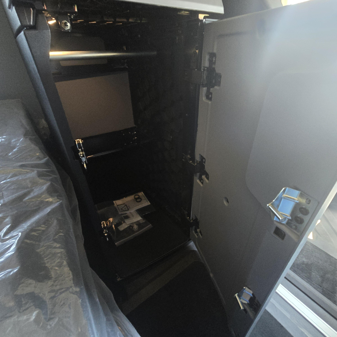 Cab interior of a new 2026 Mack PR64T76H sleeper truck showing an open side cabinet with shelves and a brochure inside; door panels and latches visible.