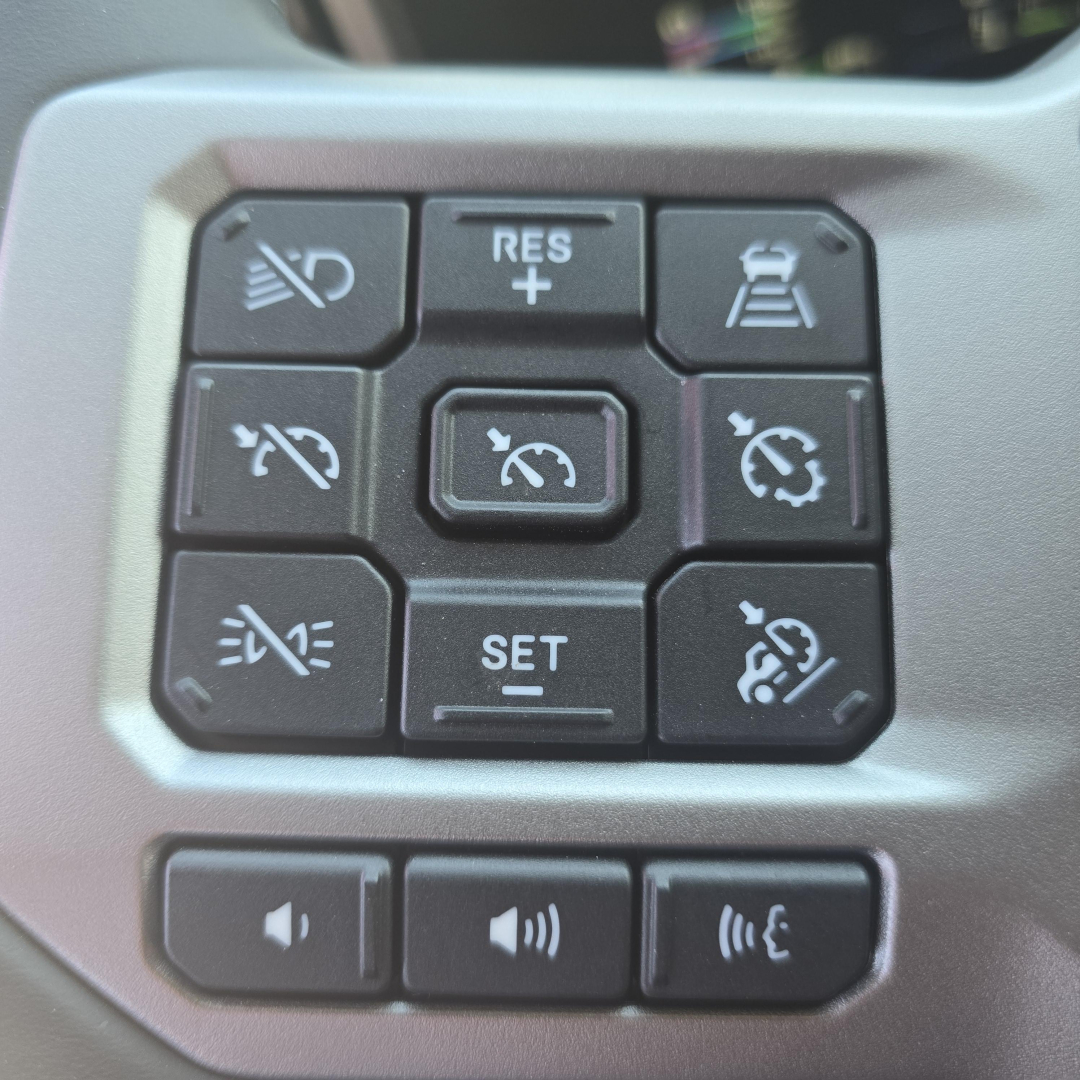 Panel of truck steering wheel controls with multiple buttons including cruise control and volume, seen in a Mack PR64T76H cockpit.
