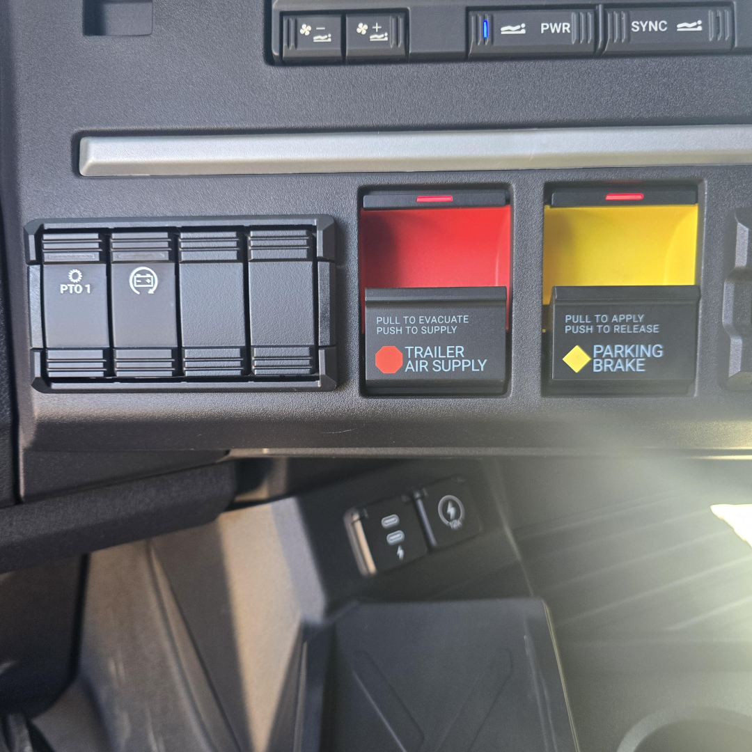 Mack PR64T76H truck dashboard controls showing trailer air supply and parking brake levers with red and yellow handles and nearby switches.