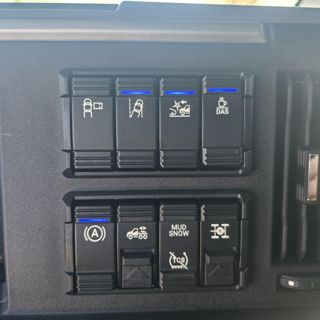 Dashboard switches panel with blue indicator lights on top row and bottom row, showing various symbols including ignition A, traction control, mud/snow, and stability features in a Mack truck interior.