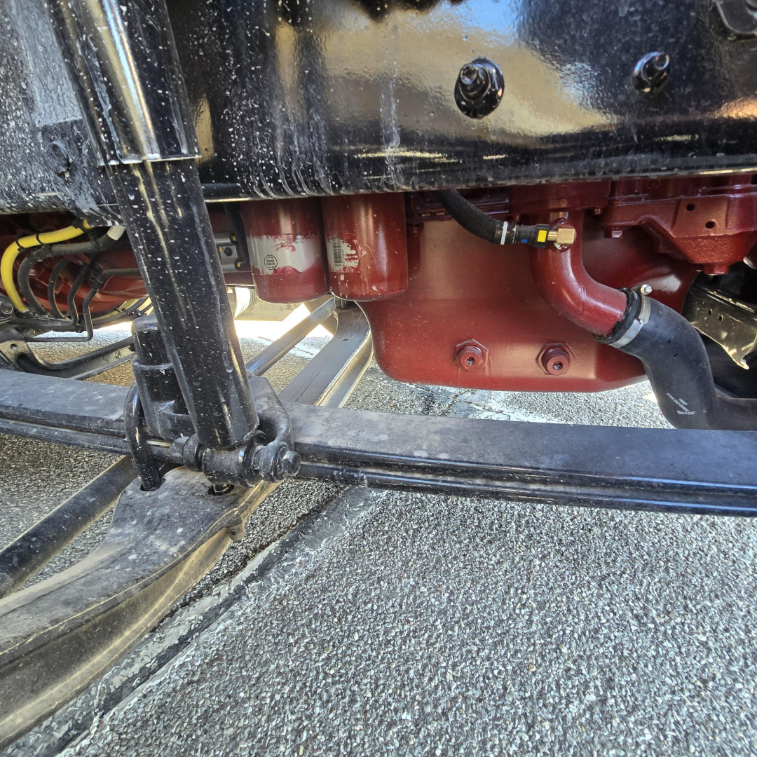 Front axle and suspension of a Mack truck with red differential housing and black frame. Visible components include a red differential/transmission housing with bolts, hoses, and a black drive shaft/axle.