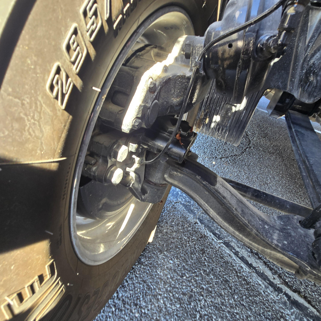 Truck front axle with tire and suspension components visible showing wheel hub and brake assembly.