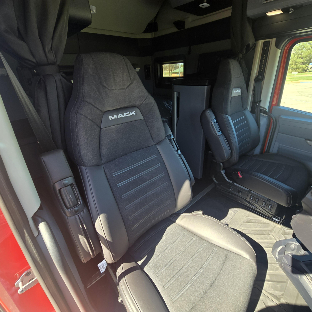 Interior cab view of a 2026 Mack PR64T76H truck showing driver and passenger seats with black fabric upholstery and 'MACK' logo on headrests.