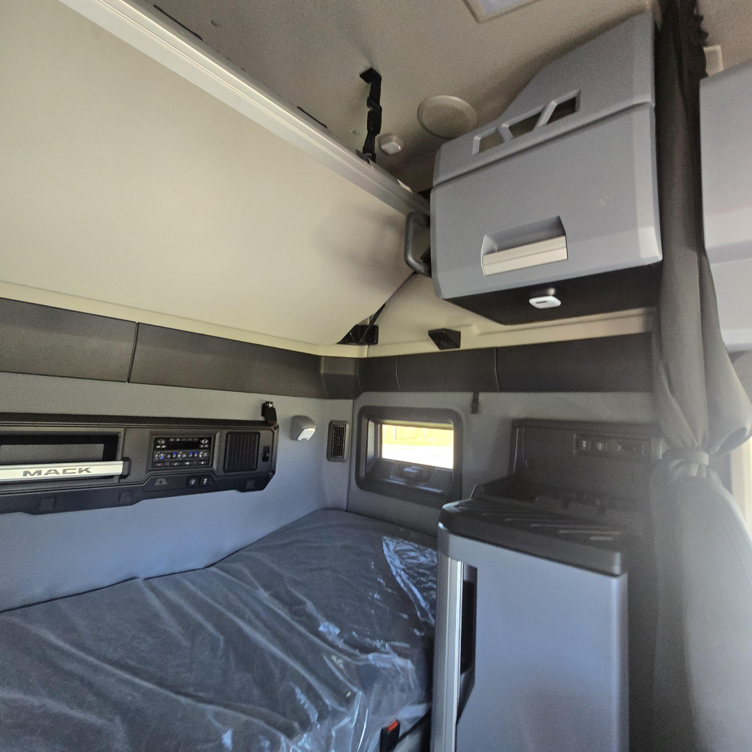 Interior sleeper cab of a Mack truck showing overhead storage and control panel with a bed and window.