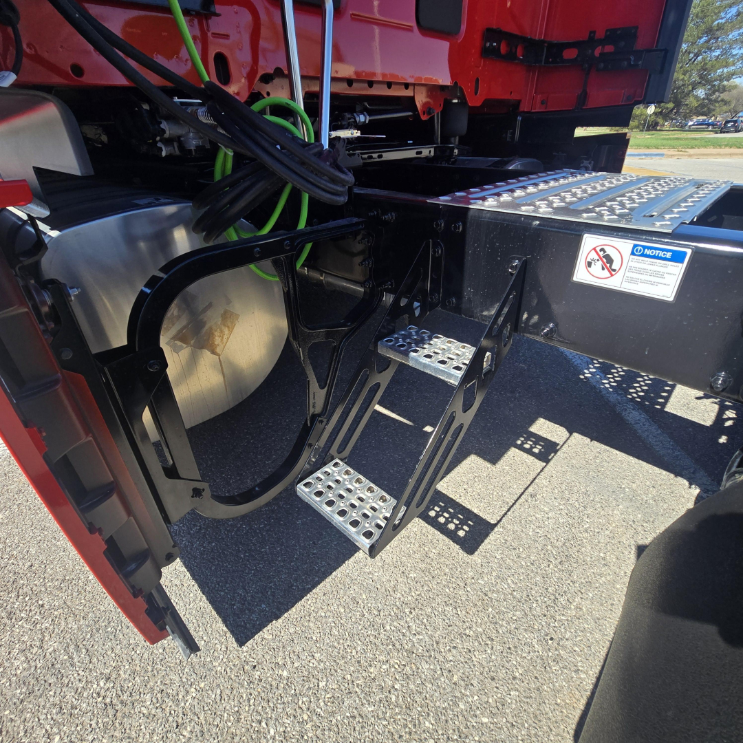 Truck side under-step area of a Mack PR64T76H showing metal perforated steps, fuel/oil reservoir, and wiring harness with labels.