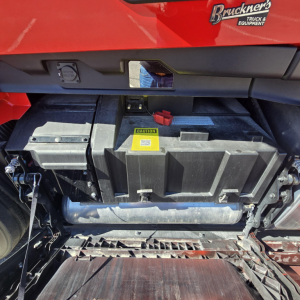 Truck engine bay showing battery box with yellow caution label and red positive terminal cover in a Mack PR64T76H.