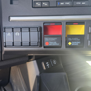 Mack PR64T76H truck dashboard controls showing trailer air supply and parking brake levers with red and yellow handles and nearby switches.
