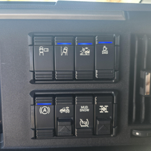 Dashboard switches panel with blue indicator lights on top row and bottom row, showing various symbols including ignition A, traction control, mud/snow, and stability features in a Mack truck interior.