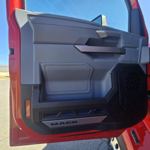 Door interior of a Mack truck cab showing panels, storage pocket, window crank area, and 'MACk' logo on the lower kick panel.