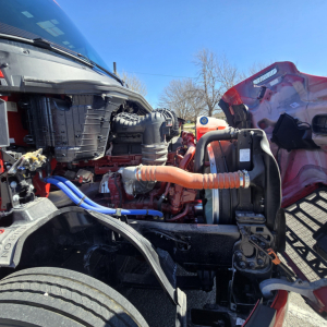 Mack PR64T76H engine bay view showing orange coolant hose, blue hose, and engine components in a truck with red hood raised.