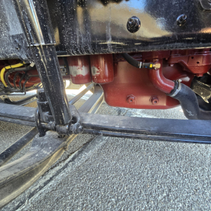 Front axle and suspension of a Mack truck with red differential housing and black frame. Visible components include a red differential/transmission housing with bolts, hoses, and a black drive shaft/axle.