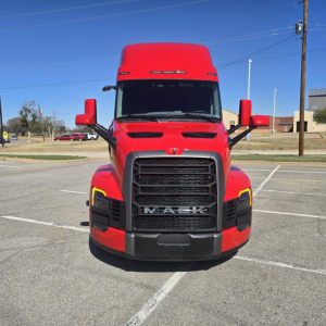 Front view of a red 2026 Mack PR64T76H truck showing grille, headlights, and large black bumper in a parking lot.