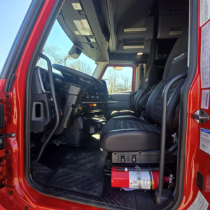 Cab interior of a 2026 Mack PR64T76H truck showing driver and passenger seats, dashboard, controls, and a fire extinguisher near the doorway.