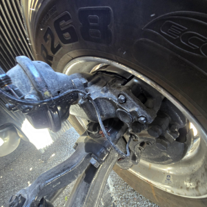 Truck rear suspension and axle assembly visible with wheel and tire showing part of the chassis and leaf spring; hydraulic lines and wiring near the hub.