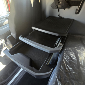 Cab interior showing open storage drawers in a new Mack PR64T76H truck sleeper with labeled compartments and a plastic organizer tray.