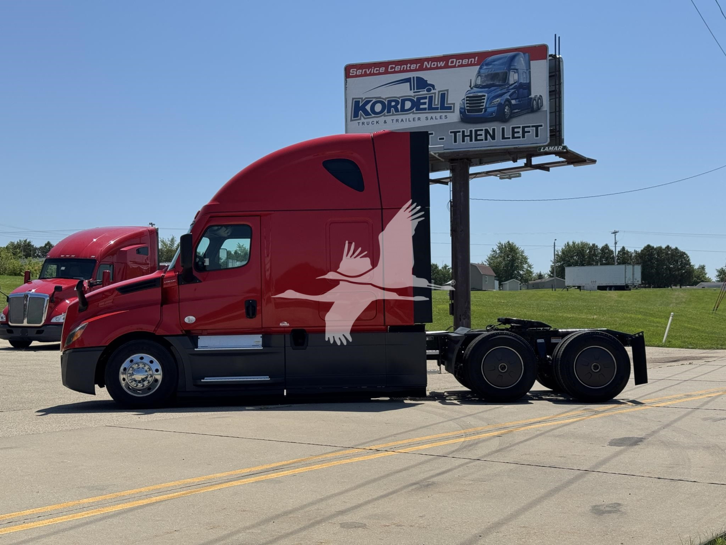 Truck side view of a red Freightliner Cascadia 126 tractor with sleeper cab; billboard behind shows hotel logo.