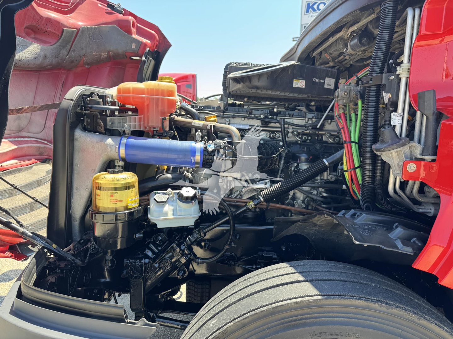 Truck engine bay showing auxiliary reservoir, blue hose with metal clamps, yellow oil cap, orange cap, black hoses, and various wiring harnesses near the radiator and frame of a red Freightliner Cascadia truck.