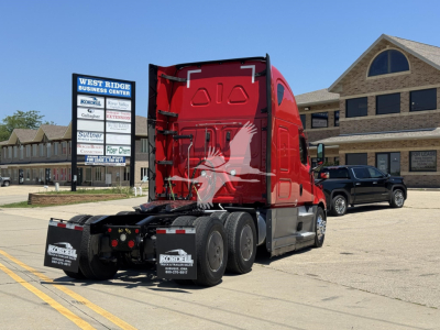 Truck tractor rear and cab of a red 2023 Freightliner Cascadia 126 in a dealership lot with a building in the background.