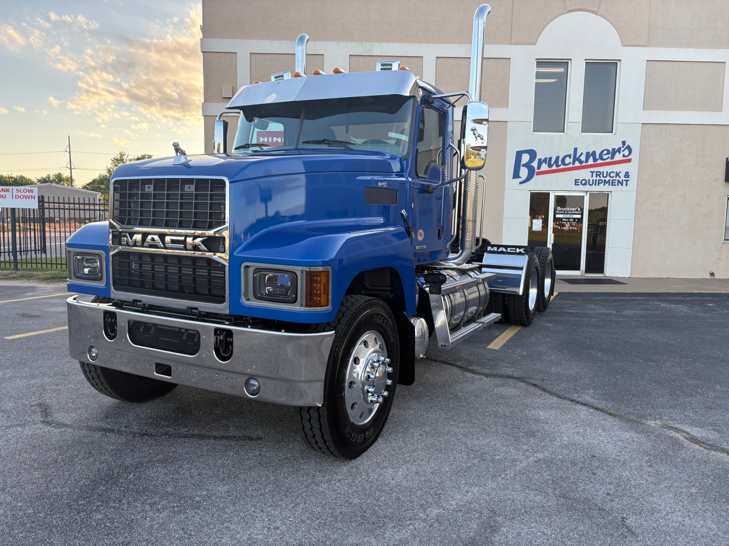 Front-left view of a blue 2026 Mack PI64T conventional daycab truck with chrome bumper, dual vertical exhaust stacks, and large grille; visible Bruckner's Truck sign in background.