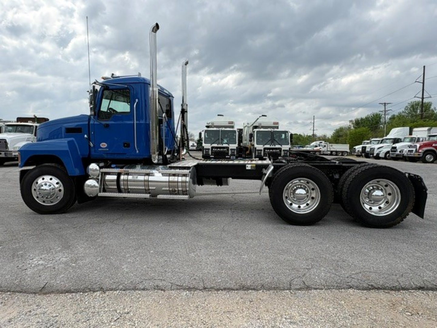 2026 Mack PI64T conventional daycab truck chassis shown in blue with dual rear axles and fuel tanks. Side view with skyline and other trucks in background.
