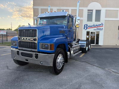 Front-left view of a blue 2026 Mack PI64T conventional daycab truck with chrome bumper, dual vertical exhaust stacks, and large grille; visible Bruckner's Truck sign in background.