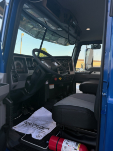 Cab interior of a 2026 Mack PI64T truck showing steering wheel, dashboard gauges, driver seat, and front passenger area; visible window, mirrors, and a floor mat.