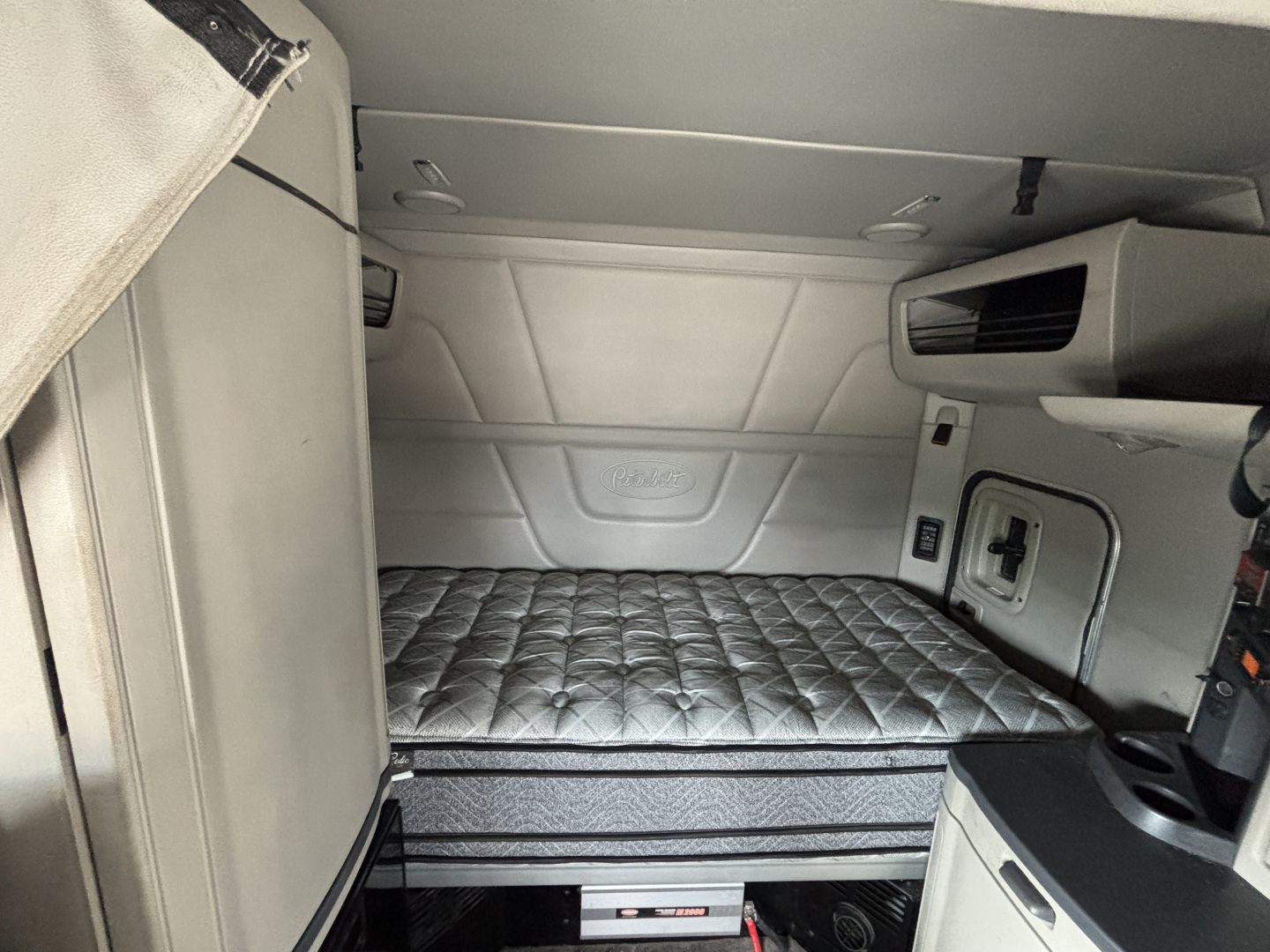 Truck sleeper cab interior with raised quilted mattress platform in a Peterbilt 579, side storage compartment, small window, and control panels visible.
