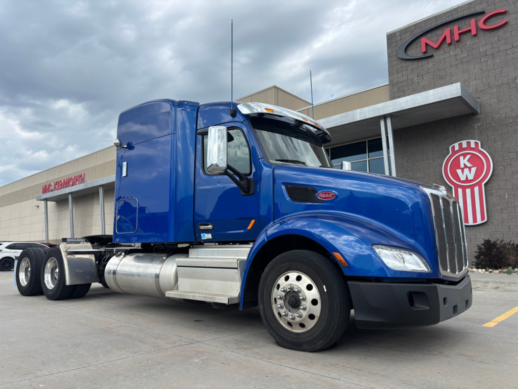 Peterbilt 579 conventional sleeper truck in blue, side view, parked in lot with KW sign in background.
