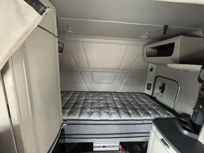 Truck sleeper cab interior with raised quilted mattress platform in a Peterbilt 579, side storage compartment, small window, and control panels visible.