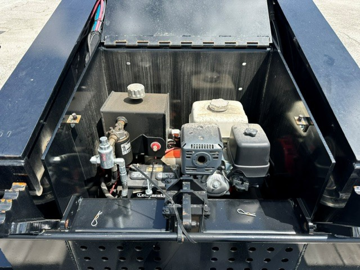 Lowboy trailer deck engine bay with hydraulic pump and control components visible, small gasoline engine, various hoses and mounting brackets.