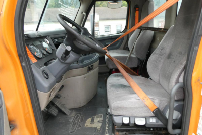Interior of a Freightliner Cascadia 125 cab showing steering wheel, dashboard, two seats, and orange seatbelt across driver and passenger seats.