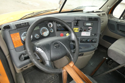 Truck cab interior showing steering wheel, dashboard gauges, and center console with warning labels and control switches.